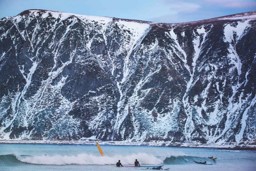 Surfere i vannet foran høyt snøkledd fjell på Flakstad i Lofoten
